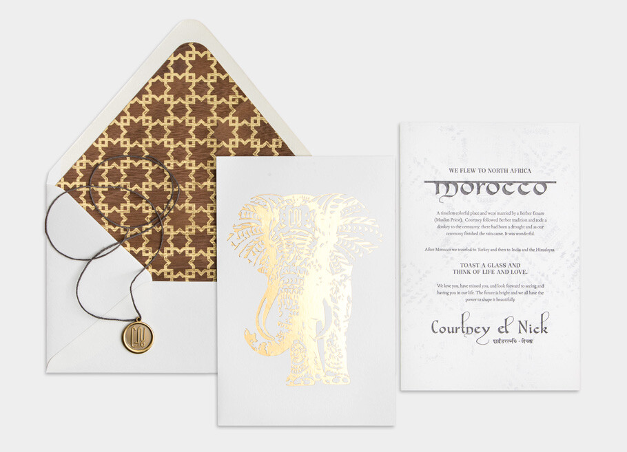 Luxury wedding invitation suite with gold foil elephant illustration, Moroccan-inspired patterned envelope liner, elegant white stationery, and wax seal detail with hanging cord on a soft gray background.