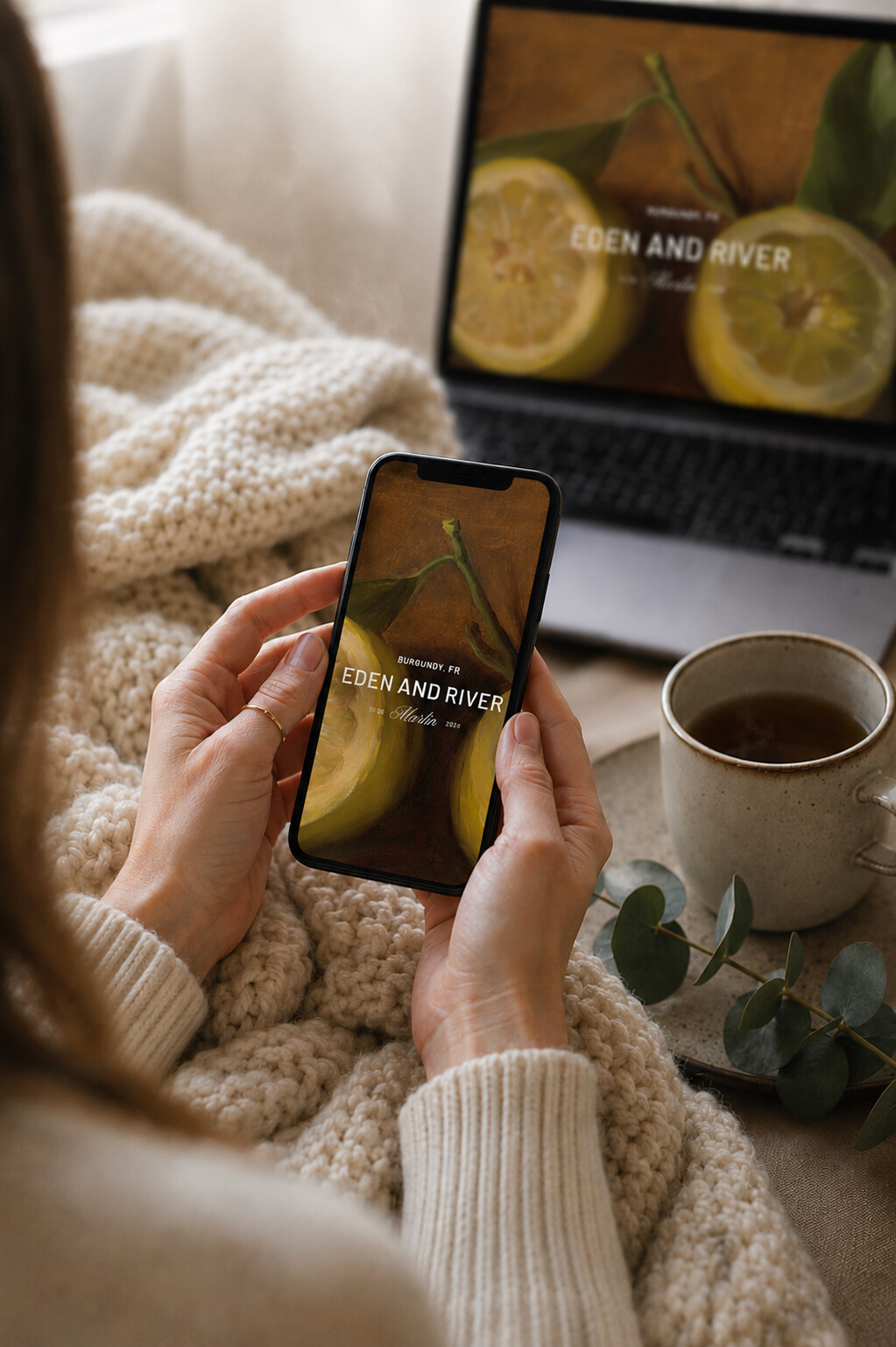 Hands holding a phone showing an elegant Bliss & Bone wedding website with the couple's names and engagement photo, beside a knit throw and tea, as a modern engagement announcement