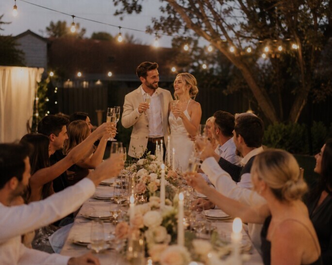 Elegant outdoor engagement party at dusk with string lights overhead, a long candlelit dinner table with floral centerpieces, and guests raising champagne glasses in a warm backyard celebration.
