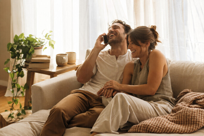 Newly engaged couple laughing on a linen sofa while telling family by phone, engagement ring catching the morning light in a sunlit apartment, Bliss & Bone