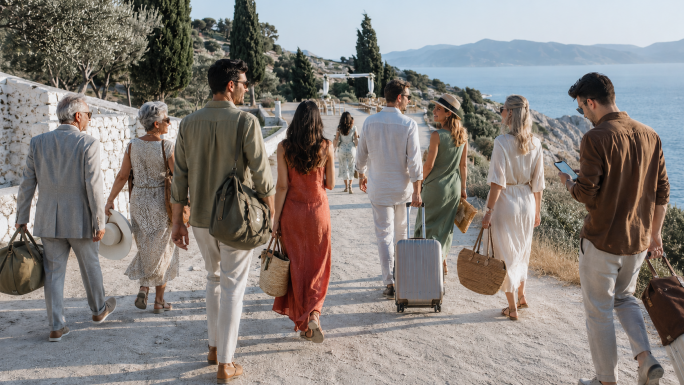 Wedding guests of mixed ages walking with weekender bags along a coastal path toward an open-air destination wedding on a seaside bluff.