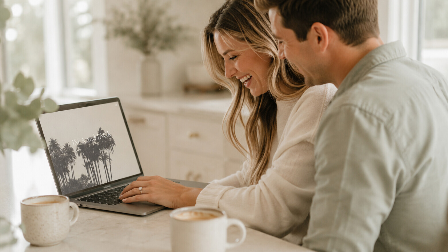 Newly engaged couple at home browsing a Bliss & Bone wedding website on a laptop, a solitaire engagement ring on the left hand, in soft natural light and a neutral greige kitchen.